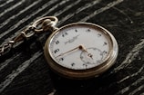 A vintage pocket watch with a white face and black numbers rests on a dark, textured surface. The watch features ornate hour and minute hands, along with a smaller subdial and a decorative chain loop.