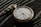 A vintage pocket watch with a white face and black numbers rests on a dark, textured surface. The watch features ornate hour and minute hands, along with a smaller subdial and a decorative chain loop.