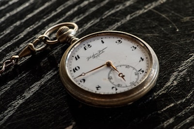 A vintage pocket watch with a white face and black numbers rests on a dark, textured surface. The watch features ornate hour and minute hands, along with a smaller subdial and a decorative chain loop.