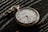 A vintage pocket watch with a white face and black numbers rests on a dark, textured surface. The watch features ornate hour and minute hands, along with a smaller subdial and a decorative chain loop.