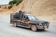 A pickup truck with a metal cage is driving along a rural road. The vehicle is carrying various items including a large tire and other objects in the back. The landscape is arid with rocky terrain and sparse vegetation. The driver is visible, wearing a hat and looking ahead.