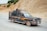 A pickup truck with a metal cage is driving along a rural road. The vehicle is carrying various items including a large tire and other objects in the back. The landscape is arid with rocky terrain and sparse vegetation. The driver is visible, wearing a hat and looking ahead.