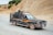 A pickup truck with a metal cage is driving along a rural road. The vehicle is carrying various items including a large tire and other objects in the back. The landscape is arid with rocky terrain and sparse vegetation. The driver is visible, wearing a hat and looking ahead.