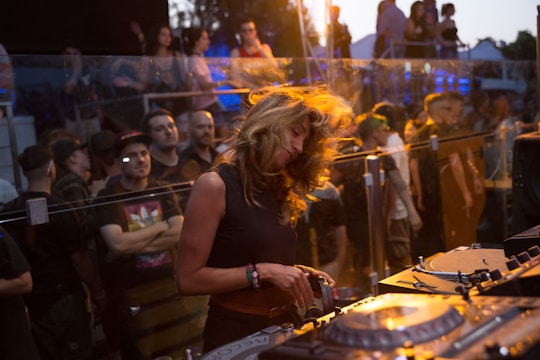 A DJ energetically playing music in front of a lively crowd. The DJ is surrounded by electronic equipment and displays a sense of immersion in the experience. The audience in the background appears engaged, with a mix of people socializing and dancing.