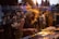 A DJ energetically playing music in front of a lively crowd. The DJ is surrounded by electronic equipment and displays a sense of immersion in the experience. The audience in the background appears engaged, with a mix of people socializing and dancing.