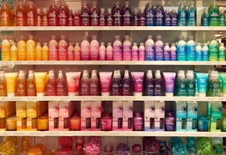 A well-organized display of colorful cosmetic or bath products arranged on multiple shelves. The bottles and boxes are in an array of colors including orange, pink, purple, blue, and green, each with unique product labeling. The arrangement creates a visually appealing rainbow effect.