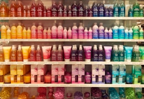 A well-organized display of colorful cosmetic or bath products arranged on multiple shelves. The bottles and boxes are in an array of colors including orange, pink, purple, blue, and green, each with unique product labeling. The arrangement creates a visually appealing rainbow effect.