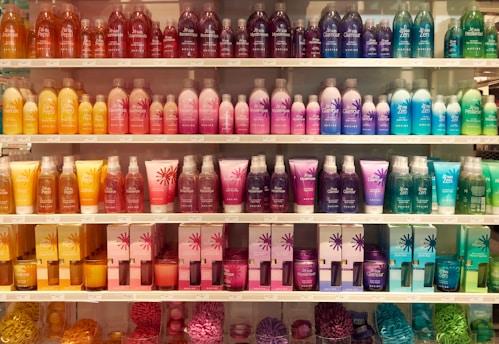 A well-organized display of colorful cosmetic or bath products arranged on multiple shelves. The bottles and boxes are in an array of colors including orange, pink, purple, blue, and green, each with unique product labeling. The arrangement creates a visually appealing rainbow effect.