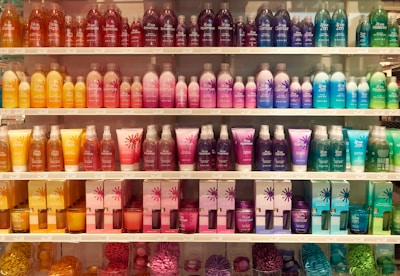 A well-organized display of colorful cosmetic or bath products arranged on multiple shelves. The bottles and boxes are in an array of colors including orange, pink, purple, blue, and green, each with unique product labeling. The arrangement creates a visually appealing rainbow effect.