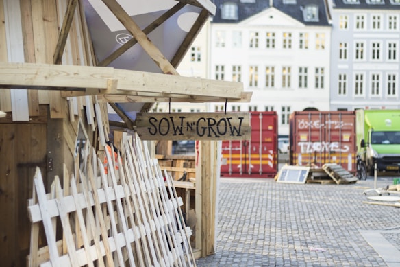 A wooden structure is adorned with a rustic sign that reads 'Sow N Grow.' Nearby are wooden picket fences leaning against the structure, and the background features cobblestone pavement and several brightly colored shipping containers.