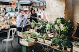 A man is spraying water on green vegetables displayed for sale at a bustling outdoor market. Various fresh produce including broccoli, greens, and other vegetables are arranged in wooden crates. Another person stands in the background, surrounded by an assortment of fruits and market items under a tent. The setting conveys a lively and vibrant atmosphere.