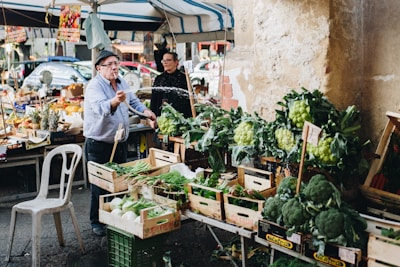 A man is spraying water on green vegetables displayed for sale at a bustling outdoor market. Various fresh produce including broccoli, greens, and other vegetables are arranged in wooden crates. Another person stands in the background, surrounded by an assortment of fruits and market items under a tent. The setting conveys a lively and vibrant atmosphere.