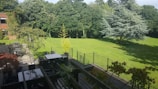 A spacious backyard features a well-maintained lawn surrounded by lush green trees. There is a patio in the foreground with dark-colored furniture, including tables and chairs. A fence separates the patio area from the lawn. The scene is bathed in sunlight, casting shadows of the trees on the grass.