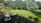 A spacious backyard features a well-maintained lawn surrounded by lush green trees. There is a patio in the foreground with dark-colored furniture, including tables and chairs. A fence separates the patio area from the lawn. The scene is bathed in sunlight, casting shadows of the trees on the grass.