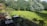 A spacious backyard features a well-maintained lawn surrounded by lush green trees. There is a patio in the foreground with dark-colored furniture, including tables and chairs. A fence separates the patio area from the lawn. The scene is bathed in sunlight, casting shadows of the trees on the grass.