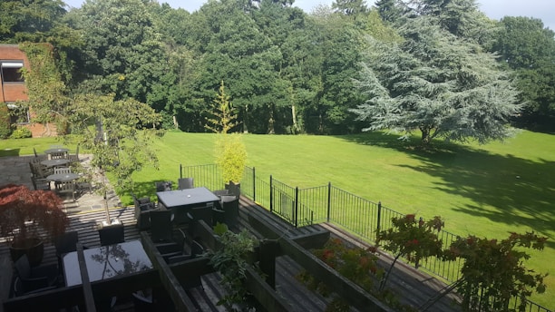 A spacious backyard features a well-maintained lawn surrounded by lush green trees. There is a patio in the foreground with dark-colored furniture, including tables and chairs. A fence separates the patio area from the lawn. The scene is bathed in sunlight, casting shadows of the trees on the grass.