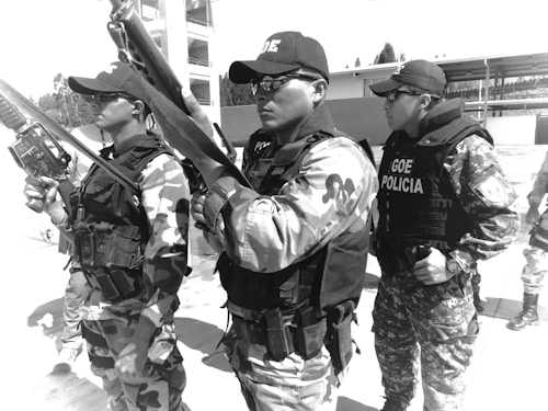 Three uniformed individuals are standing in line, wearing tactical gear and holding rifles. Their uniforms feature camouflage patterns, and they have hats and sunglasses, conveying a sense of readiness and professionalism. A building and some outdoor structures are visible in the background.