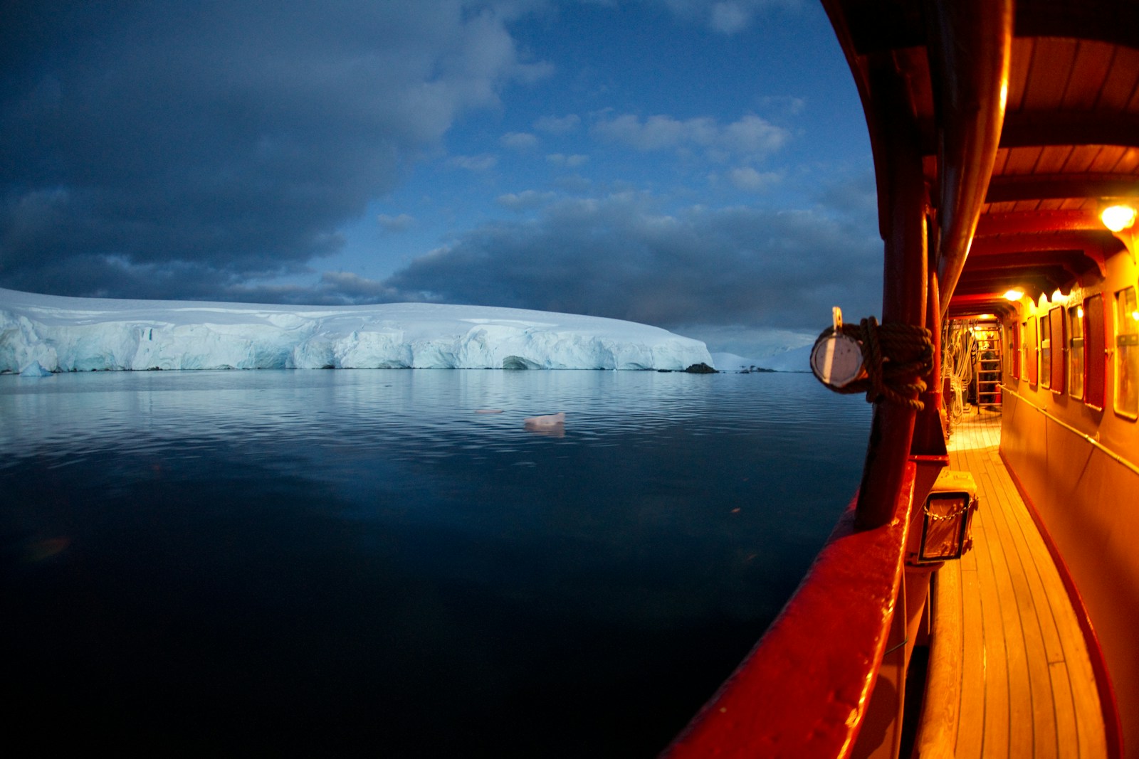 A white ship sailing dark water towards a large flat-topped iceberg on the horizon, under grey skies
