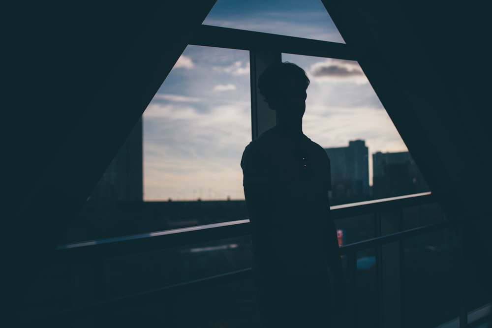 silhouette of man standing beside rails facing glass wall and buildings during blue hour