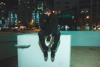 A person dressed in black sits pensively on a concrete structure in an urban setting at night. The background features glowing city lights from tall buildings, creating a moody and modern atmosphere.