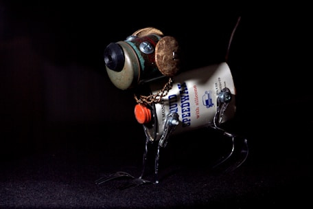 A creatively assembled sculpture appears to be a dog, constructed from various metal and mechanical parts. Its body is made from a cylindrical metal can with the word 'SPEEDWAY' printed on it. The head features a circular element resembling a camera lens, while its legs are formed from metal brackets. A chain is wrapped around the neck area, and it stands against a dark background, highlighting its unique structure.