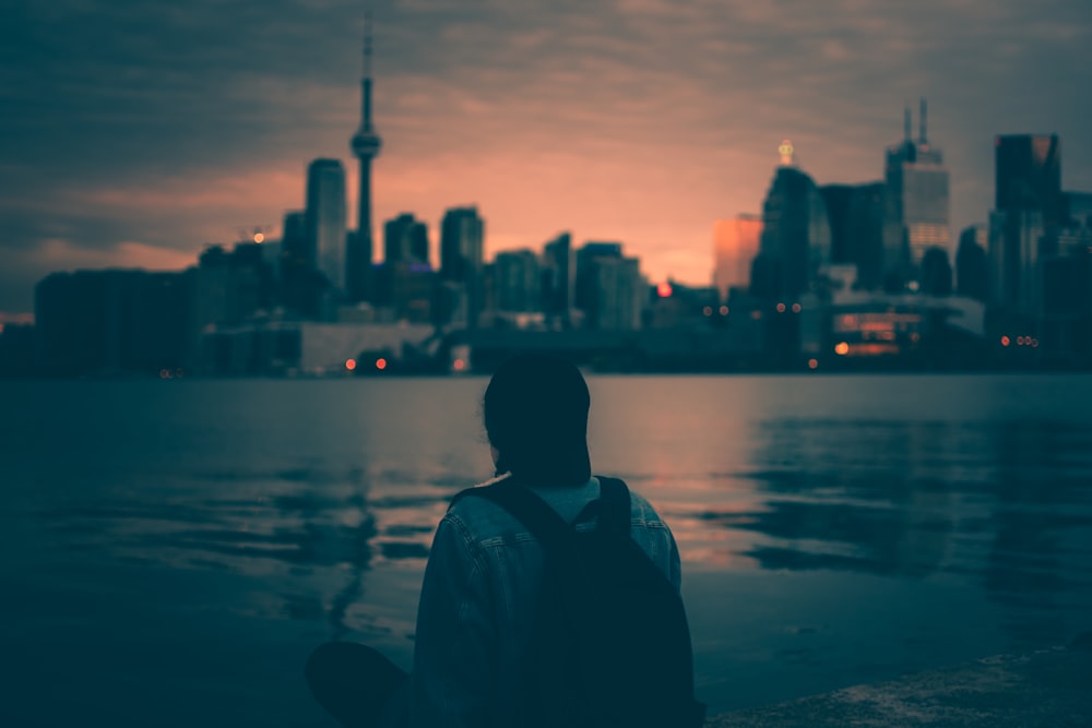 person with backpack sitting near body of water with buildings ahead