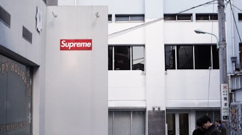A modern urban street scene featuring a building with a prominent red and white Supreme logo. The building has a minimalist design with large windows and a plain facade. There are street elements, including a street lamp and utility wires. The scene includes a few pedestrians walking by.