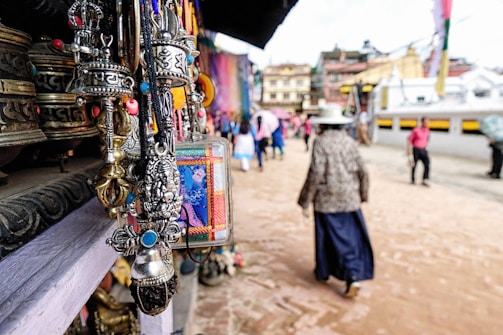 A collection of intricately designed metal pendants and ornaments is prominently displayed in the foreground. In the background, people are walking along a paved street, some wearing traditional clothing. The street is lined with buildings that have colorful facades.
