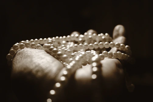 Softly lit close-up of a woman's hand gently holding a pearl necklace against champagne-colored silk fabric.