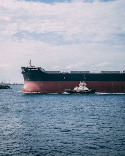A large cargo ship is navigating through a body of water, accompanied by a smaller tugboat. The sky is overcast with scattered clouds, and the vast expanse of the sea is a deep blue.