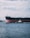 A large cargo ship is navigating through a body of water, accompanied by a smaller tugboat. The sky is overcast with scattered clouds, and the vast expanse of the sea is a deep blue.