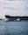 A large cargo ship is navigating through a body of water, accompanied by a smaller tugboat. The sky is overcast with scattered clouds, and the vast expanse of the sea is a deep blue.