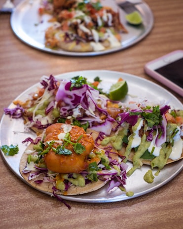 A metal plate holds two tacos topped with colorful ingredients including purple cabbage, green avocado sauce, cilantro, and a fried item. A slice of lime is visible in the background, and part of a smartphone is seen on the table.