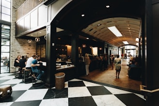A spacious indoor scene featuring a modern cafe with an open seating area. The area is filled with people enjoying coffee and conversation, with a mix of seating arrangements including bar stools and couches. Natural light streams in from large windows, highlighting the checkered floor and brick walls. The cafe counter is visible, with several customers lined up for service.