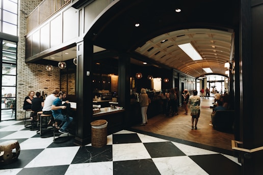 A spacious indoor scene featuring a modern cafe with an open seating area. The area is filled with people enjoying coffee and conversation, with a mix of seating arrangements including bar stools and couches. Natural light streams in from large windows, highlighting the checkered floor and brick walls. The cafe counter is visible, with several customers lined up for service.