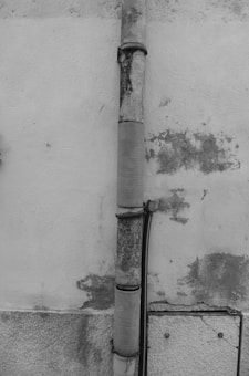 An old, weathered downpipe attached to a wall, which has patches of peeling paint and discoloration. The wall features a rough, textured surface with noticeable wear and tear. There is a smaller black cable running parallel to the downpipe, secured at various points.