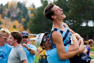 A group of young men are outdoors, smiling and laughing. One person is being lifted playfully by another, both appearing joyful and energetic. They seem to be part of a sports team, as indicated by their athletic clothing. In the background, the autumn leaves and distant houses provide a lively and colorful setting.