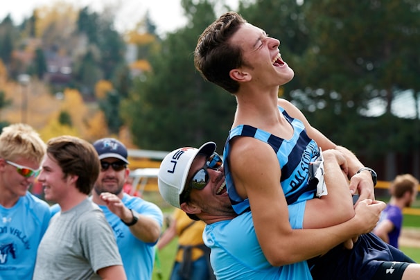 A group of young men are outdoors, smiling and laughing. One person is being lifted playfully by another, both appearing joyful and energetic. They seem to be part of a sports team, as indicated by their athletic clothing. In the background, the autumn leaves and distant houses provide a lively and colorful setting.