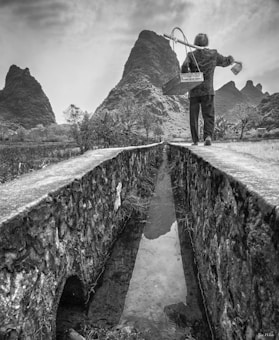 A solitary figure walks along a narrow stone pathway beside a reflective water channel. The background features striking, rugged mountain peaks, surrounded by sparse vegetation. The scene is captured in black and white, emphasizing texture and contrast.