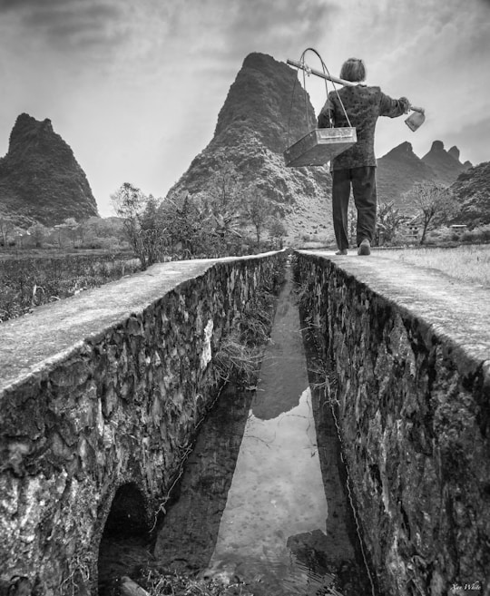 A solitary figure walks along a narrow stone pathway beside a reflective water channel. The background features striking, rugged mountain peaks, surrounded by sparse vegetation. The scene is captured in black and white, emphasizing texture and contrast.