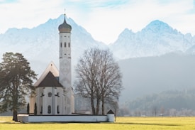 A picturesque church stands in an open field with a backdrop of towering snow-capped mountains. The church features a tall, narrow steeple topped with a bulbous spire. Leafless trees flank the building, adding a stark contrast to the white facade. The sunlight casts soft shadows, creating a tranquil and serene atmosphere.