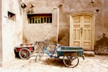 Two cargo bicycles with attached carts are positioned in front of a rustic, weathered wall and a wooden door. The bicycles are parked on a cobblestone street next to each other, one with a red cart and the other with a blue cart. The wall behind them has a gritty, aged appearance with a small rectangular window and a patchy, uneven surface.