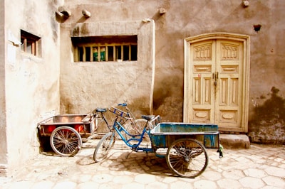 Two cargo bicycles with attached carts are positioned in front of a rustic, weathered wall and a wooden door. The bicycles are parked on a cobblestone street next to each other, one with a red cart and the other with a blue cart. The wall behind them has a gritty, aged appearance with a small rectangular window and a patchy, uneven surface.