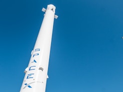 A tall, white rocket bearing the logo 'SpaceX' in blue letters is set against a clear blue sky. The rocket has aerodynamic fins near its top.