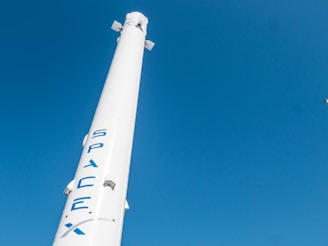 A tall, white rocket bearing the logo 'SpaceX' in blue letters is set against a clear blue sky. The rocket has aerodynamic fins near its top.