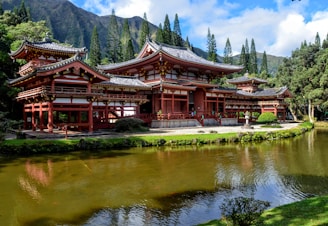 A traditional Japanese-style temple surrounded by lush greenery and set against a backdrop of tall mountains. The temple features intricate architectural details, including sweeping curved roofs with ornate eaves. A tranquil pond in the foreground reflects the structure and the surrounding landscape.