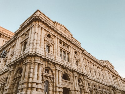 A grand, historic stone building with intricate architectural details, including carved columns and ornamental friezes. The structure has multiple arched windows and a prominent facade, showcasing classical design elements.