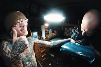 A tattoo artist is in the process of tattooing a person who appears to be enjoying the experience, giving a thumbs-up sign with a smile. The scene takes place in a dimly lit studio with various tattooing equipment around. The tattooed individual has multiple tattoos and piercings, and is wearing a beanie and glasses. The artist is focused on the work, holding a tattoo machine.