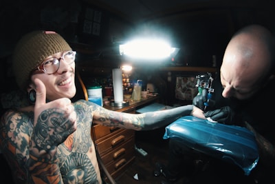 A tattoo artist is in the process of tattooing a person who appears to be enjoying the experience, giving a thumbs-up sign with a smile. The scene takes place in a dimly lit studio with various tattooing equipment around. The tattooed individual has multiple tattoos and piercings, and is wearing a beanie and glasses. The artist is focused on the work, holding a tattoo machine.