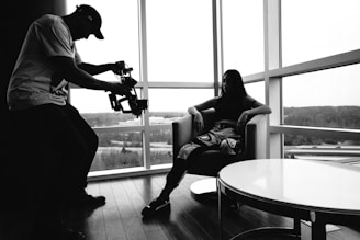 A person operates a camera rig, capturing another individual seated in a chair by large windows overlooking a landscape. The setting is indoors with a relaxed and artistic atmosphere highlighted by natural light.